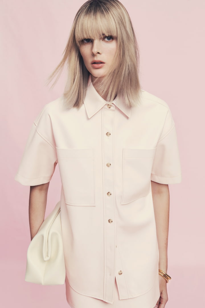 Woman in cosmetic pink PU shirt, cosmetic pink PU skirt, antique white ines pillow clutch, antique white alyssa ballet and gold noa cuff